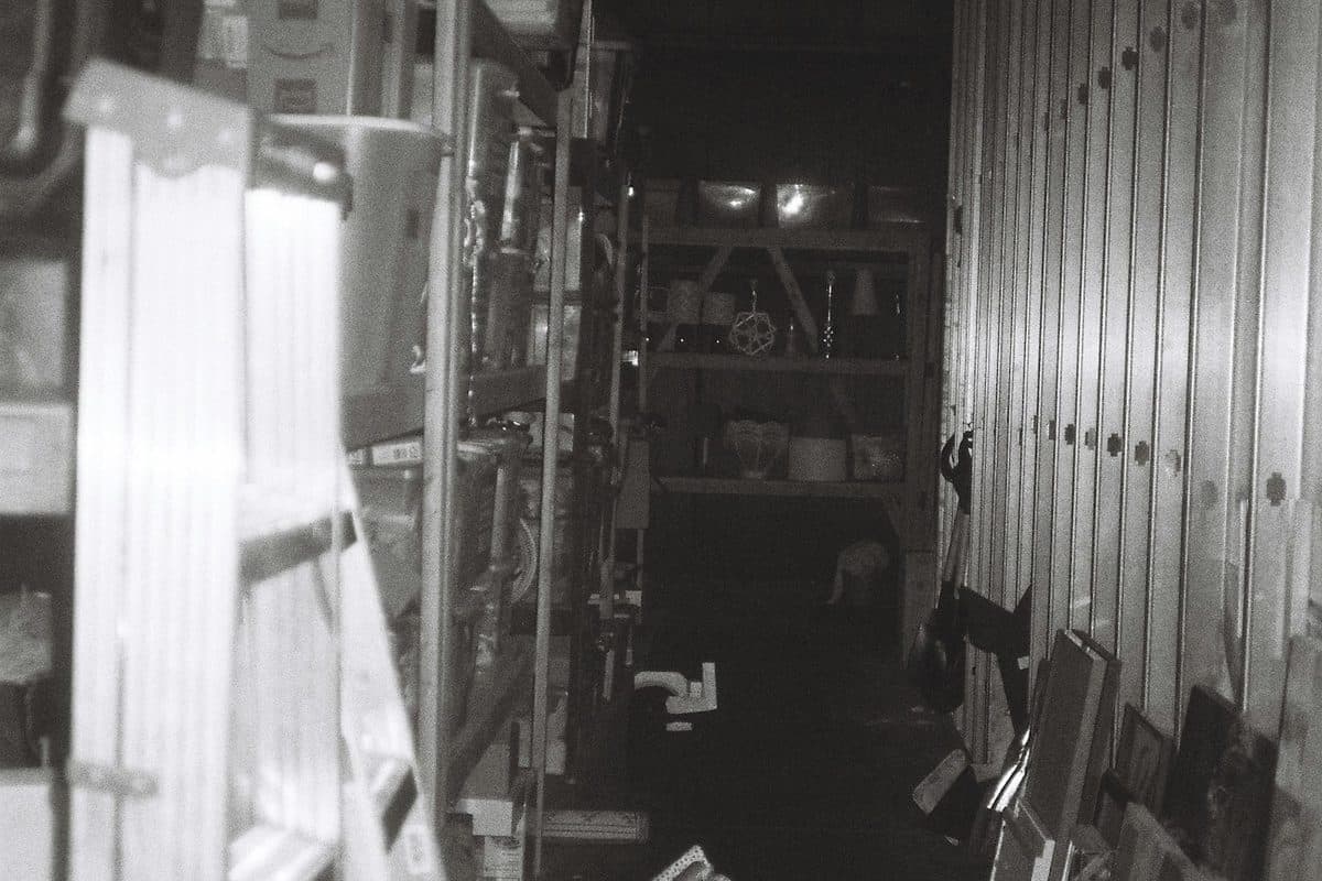 A narrow storage aisle packed with shelves, stacked bins, and a ladder, with a walking path still open