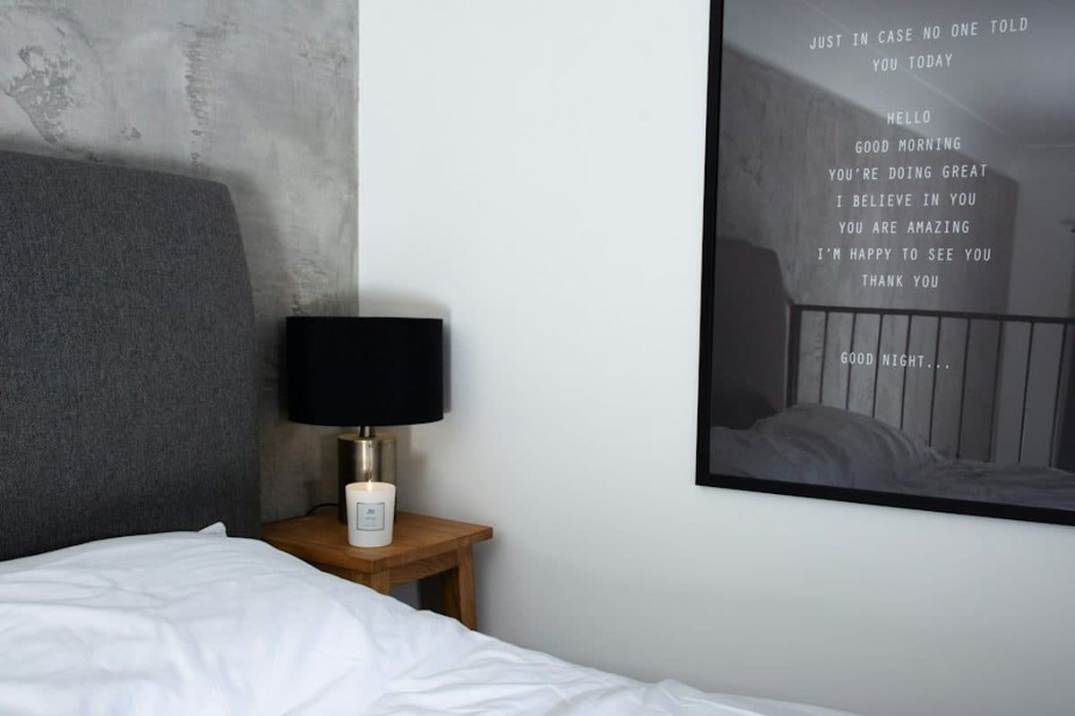 A bedroom with a gray headboard, a wood nightstand, a black lamp, a candle, and one large black frame on a mostly blank white wall