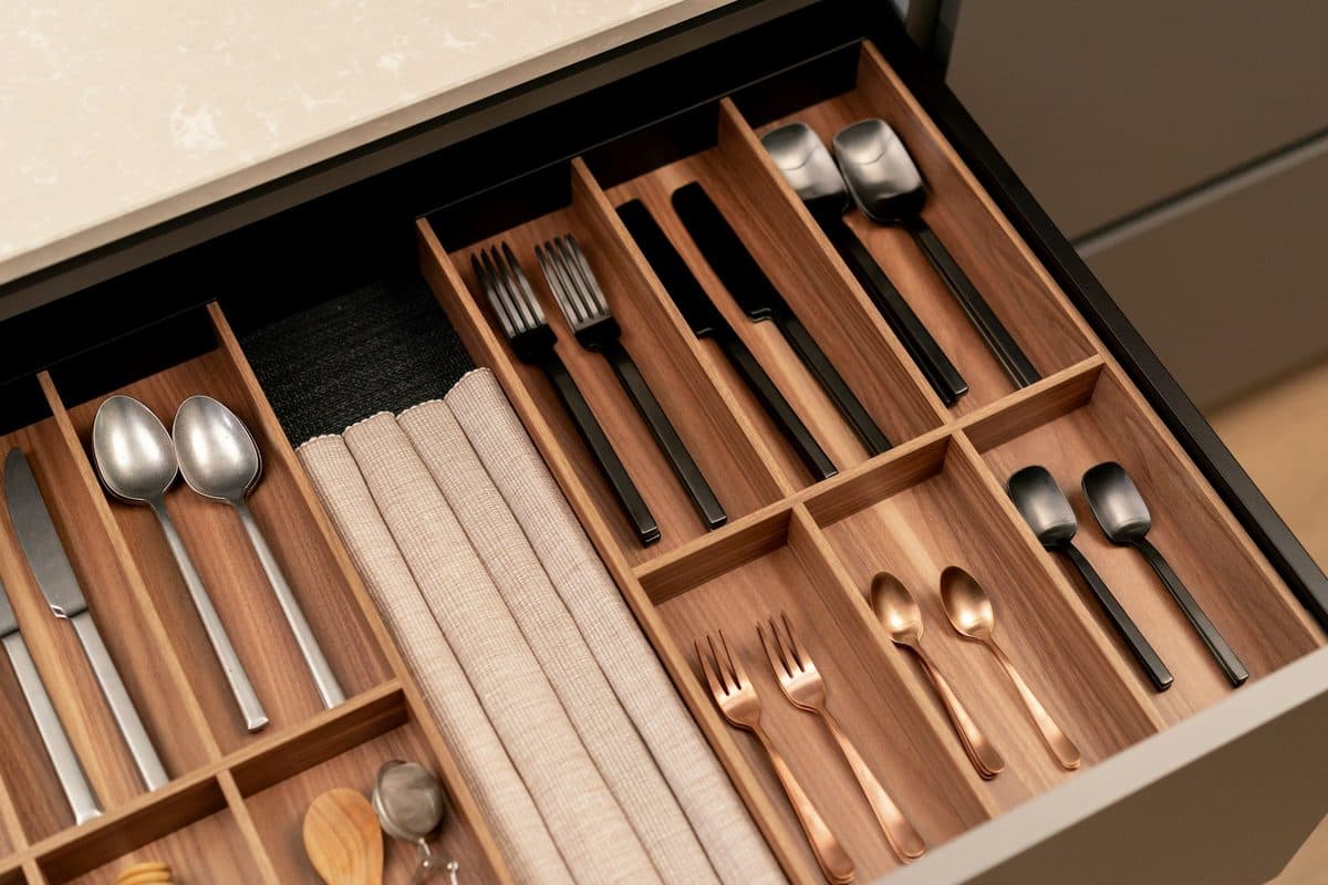 An open kitchen drawer with a rigid wood cutlery insert holding spoons, forks, knives, and rolled napkins in separate compartments