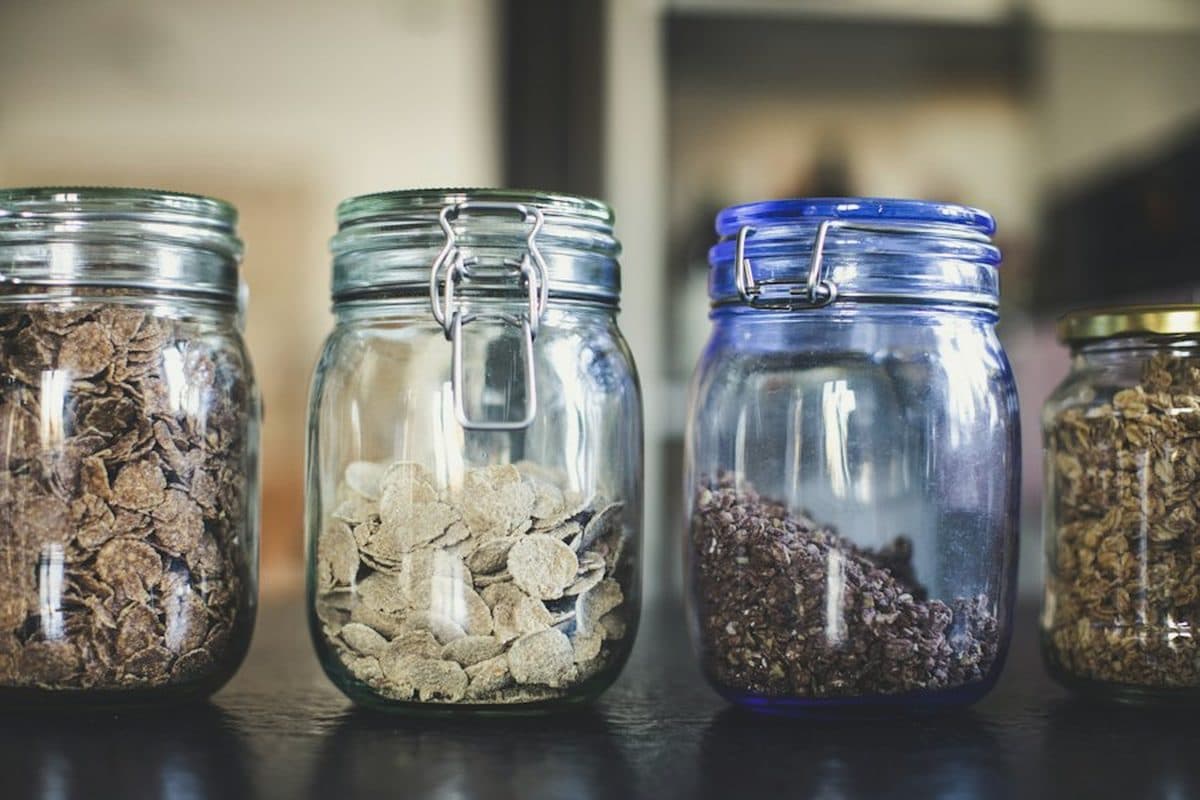 Four glass clamp-lid jars of cereal and granola in a row on a dark counter, with no handwritten labels