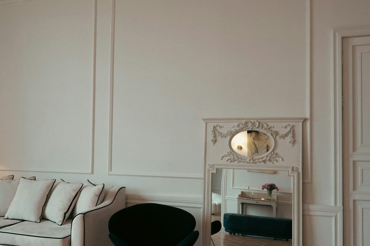 An ornate floor mirror with an arched top leaning against a white wall beside a sofa and a black chair