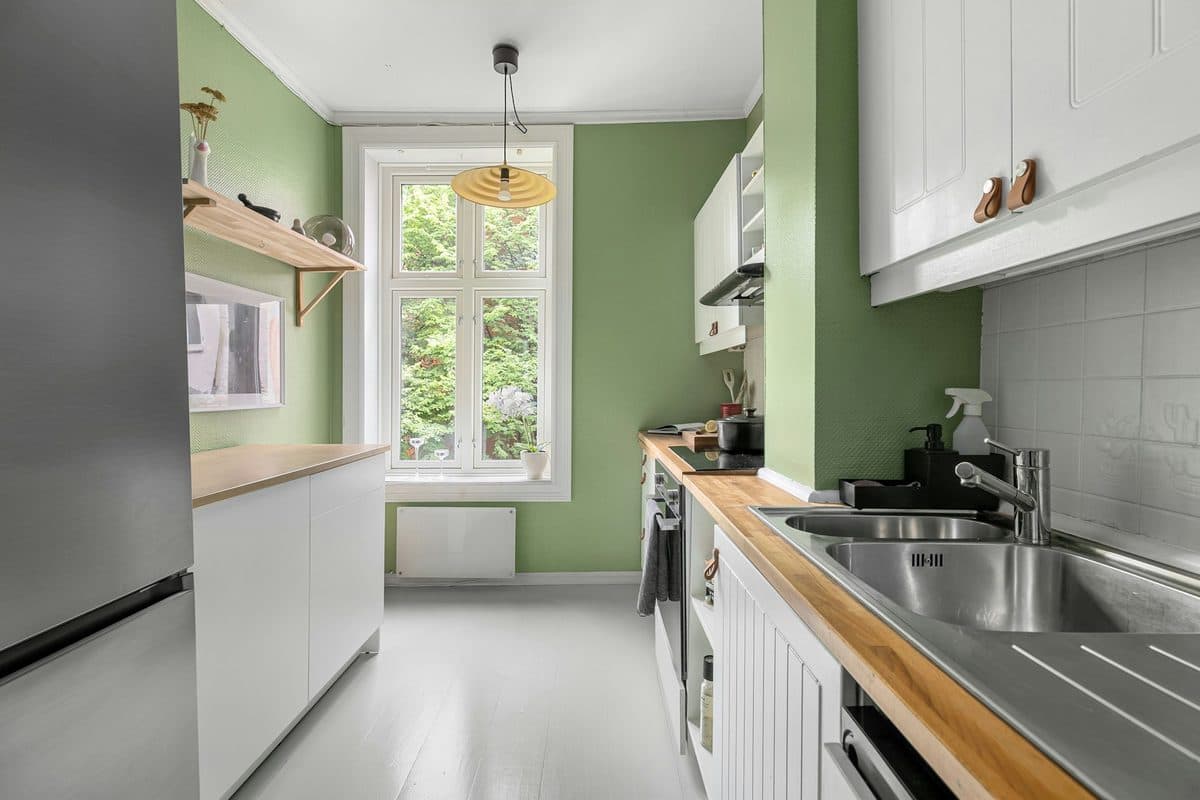 A narrow galley kitchen with white cabinets, a wood counter, a window, and a pendant light