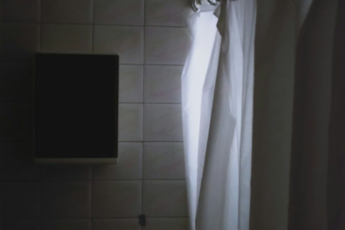A white shower curtain on metal rings against square tile, next to a dark wall cabinet