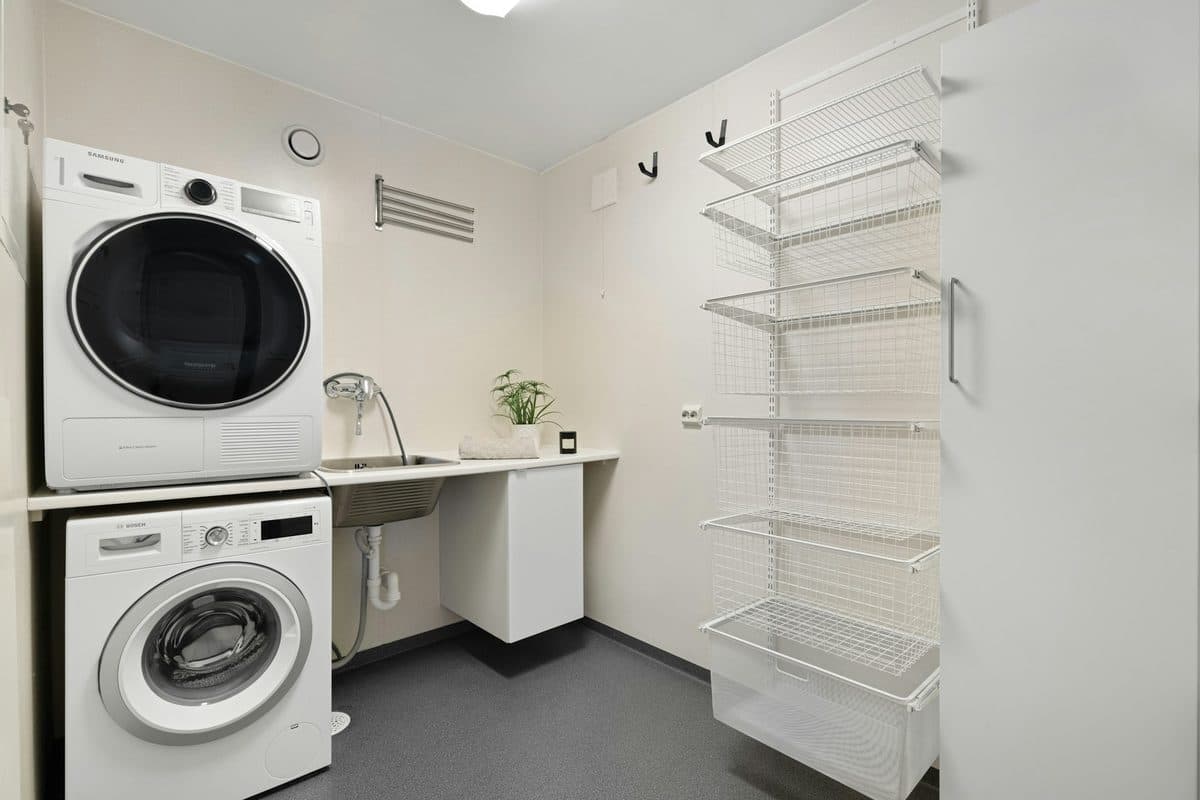 Stacked white washer and dryer beside a narrow sink, with a tall wire shelf on the opposite wall and the edge of a door at the right